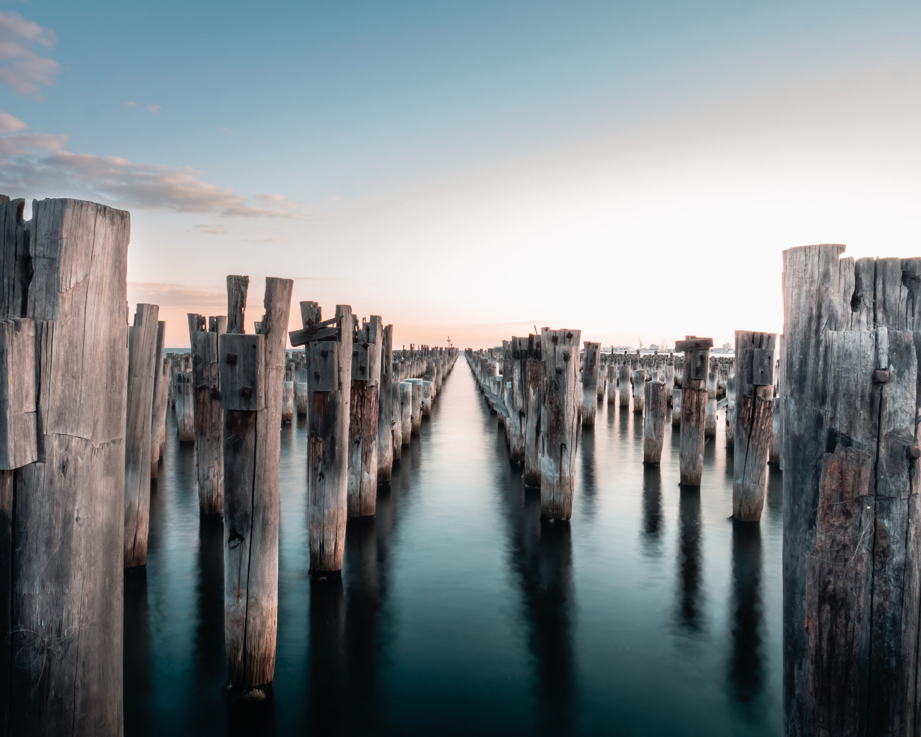 Long exposure of wooden pier posts extending into water with a soft sky in Princes Pier, Melbourne, Victoria, Australia.
